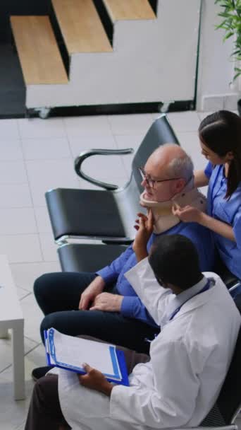 垂直ビデオ 病院のロビーで高齢者が子宮頸部の首の襟を取るのを助ける医療助手 医療治療を説明するアフリカ系アメリカ人医師 健康診断を受けた高齢者 — ストック動画