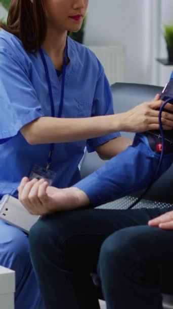 垂直ビデオ 病院の待合室で心臓検査中に高齢者の心臓パルスをチェックアシスタント 患者の血圧や高血圧を医療で測定するアジアの看護師 — ストック動画