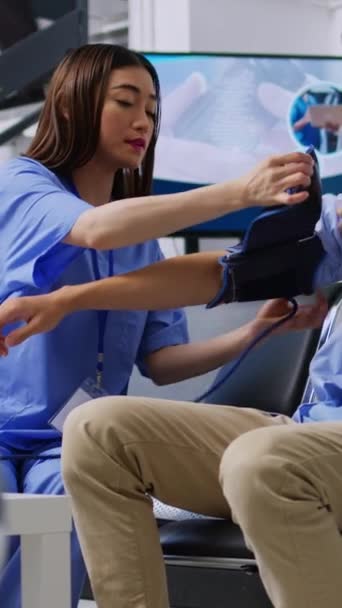 垂直ビデオ アジアの看護師は 患者の高血圧と血圧を測定し 病院の待合室で相談中に医療用計測器で心臓検査を行う アジア人男性を調べるアシスタント — ストック動画