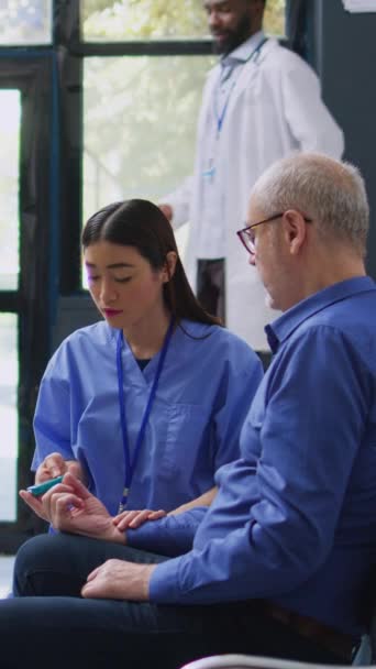 Вертикальное Видео Азиатский Ассистент Консультирует Пожилого Пациента Медицинским Глюкометром Измеряя — стоковое видео