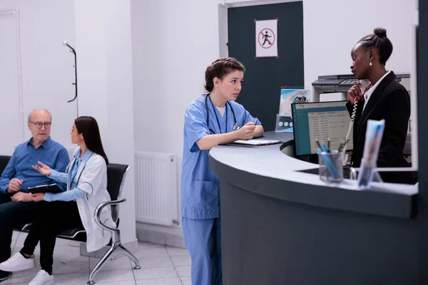 在医院候诊期间 护士站在柜台前与接待员讨论预约检查事宜 谈论健康的妇女是服务 她们努力提供高质量的护理 — 图库照片