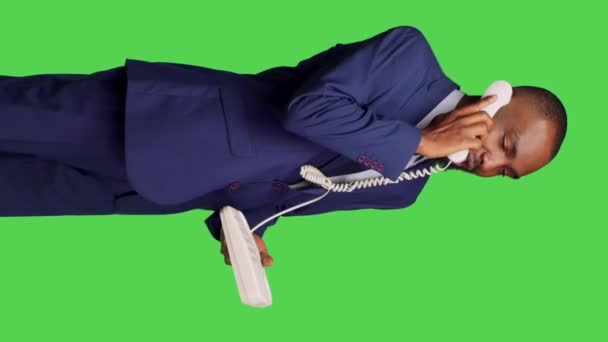 垂直ビデオ 固定電話を使ってマネージャーに電話をかけ スタジオでのリモートチャットのためのコードでオフィスの電話に応答するアフリカ系アメリカ人の男性の側面図 緑の画面の背景にポーズをとる男性従業員 — ストック動画