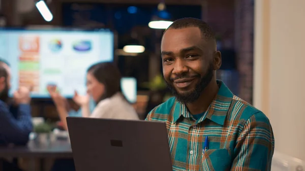 アフリカ系アメリカ人のオフィス従業員を笑顔ノートパソコンの肖像画に取り組んでいます 若い男は カメラを閉じて 会社の財務プレゼンテーション ビジネスミーティングの背景を見て コンピュータを保持 手持ち撮影 — ストック写真
