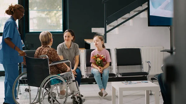 Elderly patient in wheelchair meeting with family in hospital waiting room, talking to woman and little girl in lobby. Senior person with disability receiving medical assistance at health center.