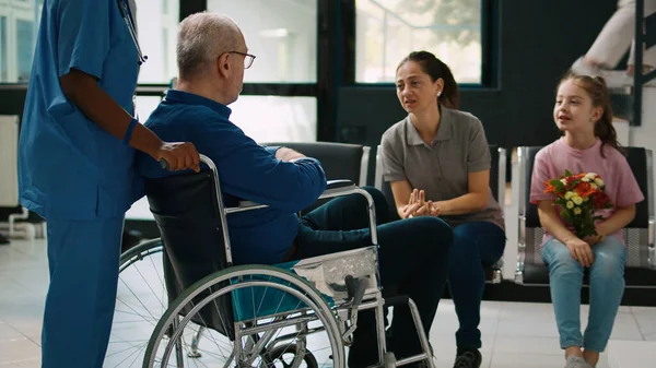 Nurse helping man in wheelchair to meet with family in clinic lobby, old patient dealing with chronic impairment at facility. Mother and child visiting senior person in waiting room.