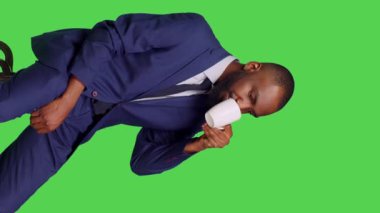 Dikey video: Sandalyede kahve içen erkek çalışanın yan görüntüsü, stüdyoda yeşil ekran arkaplanda oturuyor. Genç işadamı kafein içeceği ya da kamera önünde sıcak bir içecek içiyor.