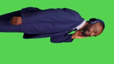 Dikey video: Genç yetişkin profili kulaklık kullanıyor ve kamera önünde müzik dinliyor, stüdyoda şarkı söyleyip dans ediyor. Erkek şirket çalışanı, ses kulaklığı takıyor ve mp3 ile mutlu.