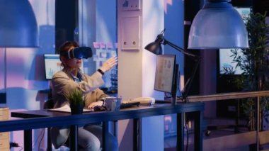 Young worker wearing virtual reality goggles in office, planning financial report to help increase profit. Business woman working late on economy graphs using vr headset at desk.