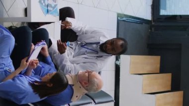 Dikey video: Doktor muayene sırasında yaşlı adama tıbbi teşhisi açıklıyor, hemşire panoya ilaç tedavisi yazıyor. Servikal tasma takan yaşlı bir hasta.