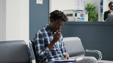 African american patient filling in checkup report papers, sitting in hospital waiting room lobby. Male person reading medical form files before attending appointment with general practitioner.