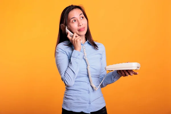 Filipino woman talking on landline phone call, using telephone with cord standing over yellow background. Cheerful model using office phone with cord having remote discussion in studio