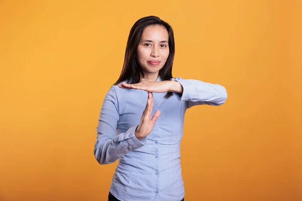 Filipino woman showing timeout and break gesture in studio, doing t shape sign with hands to pause action. Serious young adult gesticulating refuse standing over yellow background. Stop concept