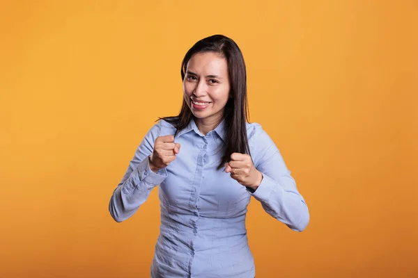 Defensive asian woman doing self defense gesture when encountering a life threat. Brave angry young adult clenching fists ready for boxing match, standing in studio over yellow background