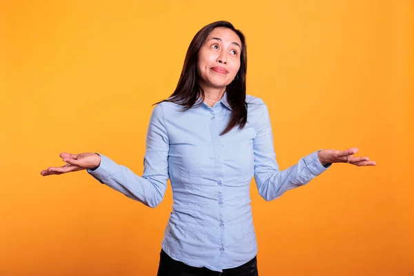 Pensive doubtful woman doing i dont know gesture, posing indecisive with shrugging shoulders in studio over yellow background. Asian young adult making confused uncertain facial expression.