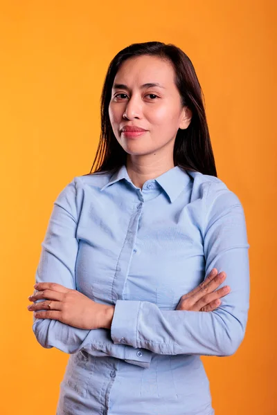 Portrait of carefree filipino woman posing with confidence, holding hands in crossed position during photoshot. Pretty model feeling attractive in studio over yellow background. Positive expression