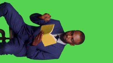 Dikey video: Stüdyoda edebiyat kitabı okuyan modern girişimcinin koltukta oturan takım elbise giymesinin ön görüntüsü. Olumlu iyimser adam yeşil ekranda ders veriyor.