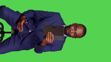 Dikey video: Erkek ofis çalışanının dijital tablette uzaktan telekonferans sohbetini kullanarak stüdyoda online video görüşmesi yaparken ön görüntüsü. Genç yetişkinler modern insanlarla video konferansında sohbet ediyorlar.