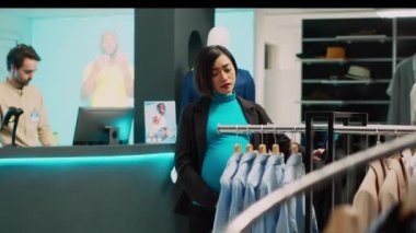 Asyalı hamile bir kadın modern kıyafetleri askıda kontrol ediyor. Müstakbel anne resmi kıyafet almak istiyor. Hamilelik göbeği olan genç bir müşteri moda mağazası ürünlerini inceliyor..