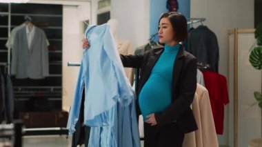 Bebek yumrusu olan Asyalı kadın moda mağazasında moda tişörtler alıyor, hamile müşteri askılardaki kıyafetlere bakıyor. Kadın müşteri elbise mağazasında alışveriş yapıyor. Üçayak atışı.