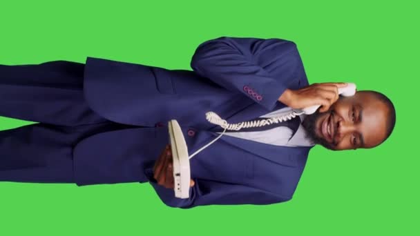 垂直ビデオ 固定電話を使用してマネージャーに電話をかけ スタジオでのリモートチャットのためのコードでオフィスの電話に応答するアフリカ系アメリカ人の男性の正面図 緑の画面の背景にポーズをとる男性従業員 — ストック動画