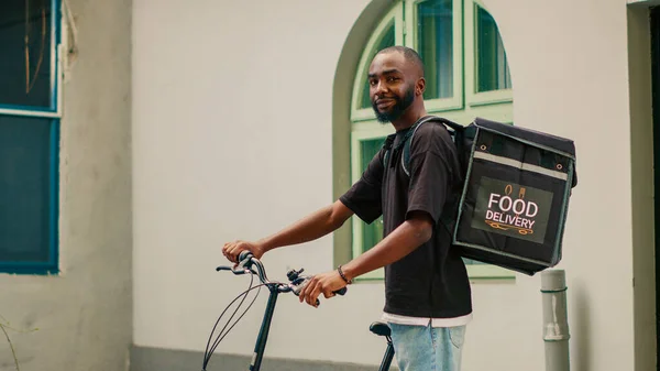 アフリカ系アメリカ人男性宅配便のお客様に食事のパッケージを与えるために自転車に乗って フロントドアでバックパックを運ぶ 自転車で働く配達人とレストランを配達する テイクアウト食品 — ストック写真