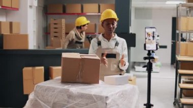 Afro-Amerikalı kadın, kutularda reklam ürünleri satıp depoda akıllı telefonla çalışıyor. Tulum giymiş kadın çalışan sosyal medya ve endüstriyel stoklar için reklam videosu çekiyor..