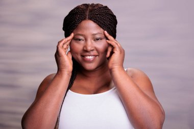 Afrika kökenli Amerikalı kadın yüz masajı yapıyor. Cilt bakımını yapıyor. Yüze parmaklarıyla dokunuyor ve kameraya bakıyor. Artı, gülümseyen güzel model taze temiz deri portresi yapıyor.