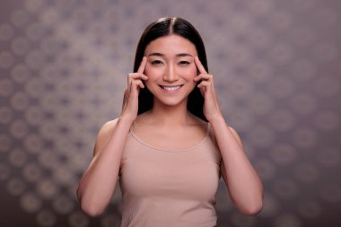 Cilt yüzlü Asyalı bir model, gözleri kırışıklık önleyici masaj prosedürünü, şakaklara dokunmayı ve gülümsemeyi gösteriyor. Doğal güzellikte genç bir kadın cilt bakımı rutini yapıyor ve kameraya bakıyor.