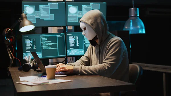 Cyber scammer wearing mask and hood to hack computer system, breaking into servers to steal big data. Masked man looking dangerous and scary, impostor creating security malware. Handheld shot.