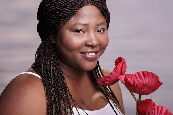 Body positive lady with red poppy flowers face portrait. Smiling african american curvy beautiful woman holding plant with red blossoms, posing in studio and looking at camera