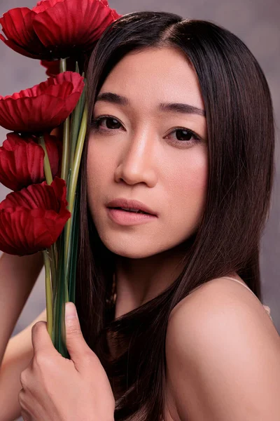 Young woman leaning red flowers to fresh hydrated skin portrait. Skincare asian model wearing natural makeup holding blooming plants with beautiful blossoms near face close up