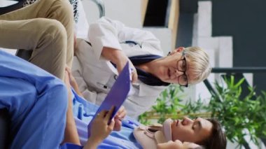 Dikey video: Sağlık görevlileri bekleme alanında muayene sırasında boyunluğu çıkarmak için yaralı bir hastayla tartışıyor. Asyalı asistan elinde pano tutuyor. Tıbbi uzmanlık yazıyor, sağlık hizmetleri planlıyor.