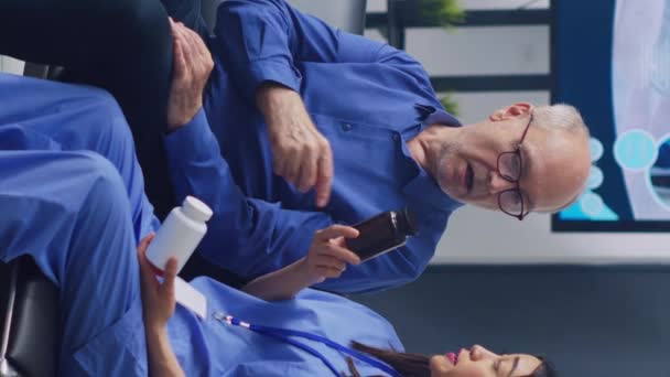 Pionowe Wideo Azjatycki Asystent Pokazuje Butelki Antybiotykami Starszemu Pacjentowi Omawiając — Wideo stockowe