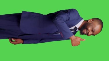 Dikey video: Kamera önünde baş parmağını gösteren ve yeşil ekran arkaplanı için onay veren erkek çalışanın yan görüntüsü. Takım elbiseli genç adam stüdyoda anlaşma jesti yapıyor, tamam.