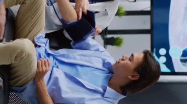 Dikey video: Hastanenin bekleme alanında yapılan tıbbi muayene sırasında hastanın yüksek ya da düşük nabzını kontrol etmek için tonometre kullanan yaşlı bir doktor. Kıdemli doktor Asyalı adamın hipertansiyonunu ölçüyor.
