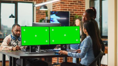 Team of diverse workers analyzing computers with greenscreen template, women looking at mockup display. Content creators using isolated chroma key with blank copyspace on monitors.