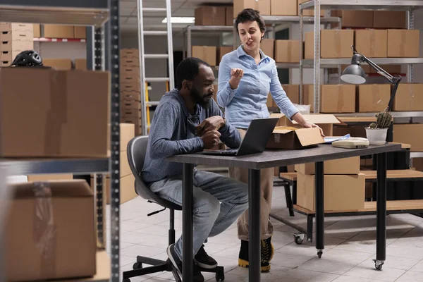 时尚工厂的员工在把服装订单放进纸盒后 分析了笔记本电脑上的客户运输数据 讨论了包装运输问题 在仓库工作的不同雇员 — 图库照片