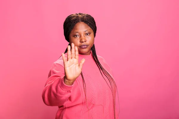 Negative woman doing stop symbol with palm expressing denial and rejection posing in studio with pink background. Young adult denying and disapproving with idea, raising hand decline suggestion.