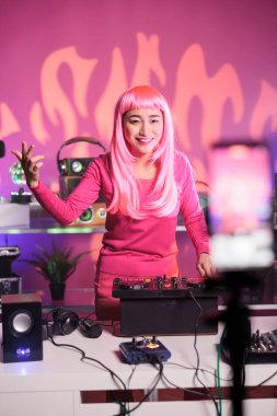 Pembe saçlı sanatçı, profesyonel mikser konsoluyla telefon kamerasını karıştırarak tekno şarkı söylerken stüdyoda eğleniyor. DJ kadın gece kulübünde performans sergiliyor.