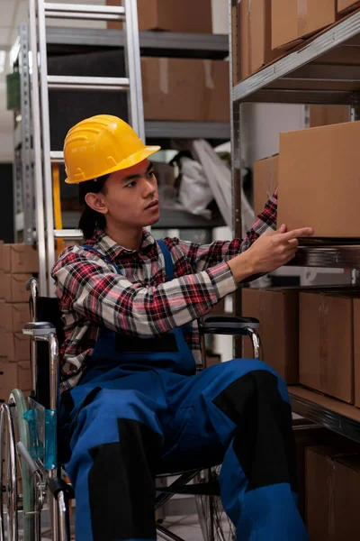 Asian shipment manager with disability picking order in industrial storehouse. Postal service warehouse employee putting cardboard box on shelf, working in inclusive workplace
