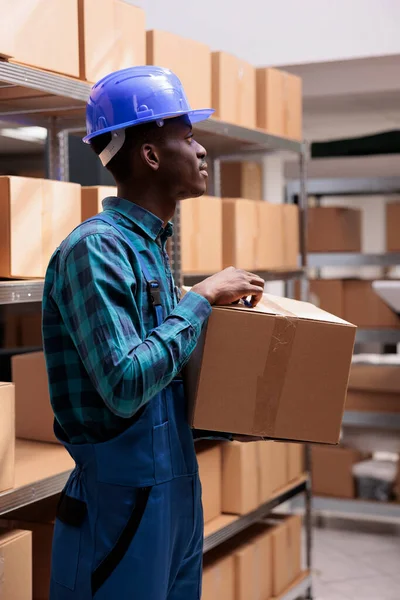 Warehouse worker with carton managing parcels receiving and dispatching process. Young african american man wearing blue unifrom and helmet working in goods distribution storehouse