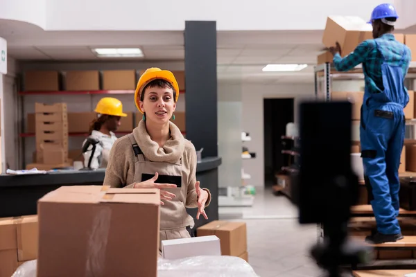 仓库工人记录智能手机视频与库存管理解释航运公司的实习生 在仓库里笑着的年轻女人和在手机摄像机前说话的头盔 — 图库照片