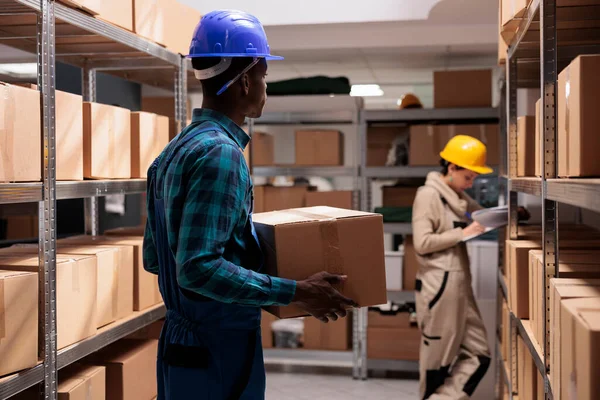 工业仓库助理准备纸板箱待运 站在仓库过道上 头戴防护帽 头戴货物包的非裔美国人 — 图库照片