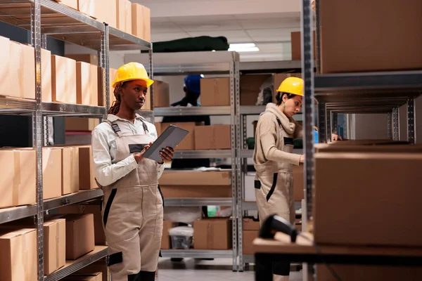 仓库经理监督货物库存和审核申请 非洲裔美国妇女站在纸板箱架上的仓库里 使用数字平板电脑 — 图库照片