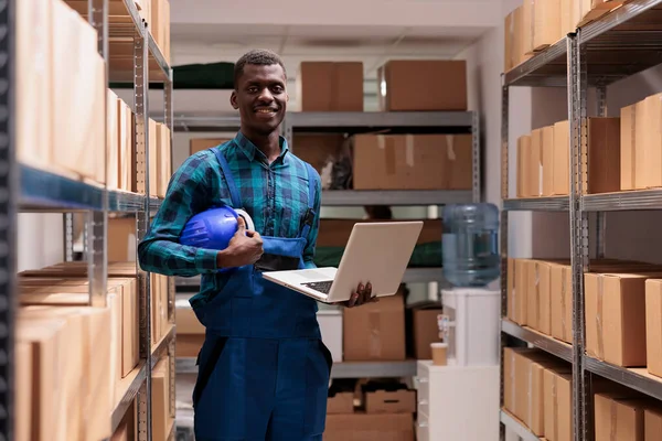 African american distribution center manager holding laptop in warehouse portrait. Smiling delivery service storage operator wearing protective uniform and helmet standing near shelf with containers