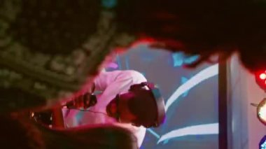 Dikey video: Sahnede yetişkin bir erkek müzik istasyonu kullanıyor, diskotekte müziği karıştırıyor, bir grup arkadaş parti yapıyor ve dans pistinde zıplıyor. Elektronik ritimlerde dans eden genç mutlu insanlar. El kamerasıyla.