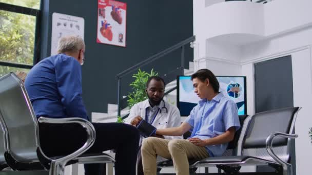 医師は アジアの患者の高血圧を測定するために 血圧や脈圧をチェックします アフリカ系アメリカ人医師が病院のロビーで検査をしています 疾患診断 — ストック動画