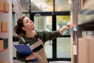 Depo çalışanı karton kutularla dolu raflara bakıyor. Ürünleri göndermeye başlamadan önce müşterilerin siparişlerini hazırlıyor. Depo yöneticisi eşofmandan giydiği üniformayla nakliyatı onaylıyor.