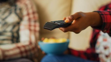 Woman switching channels with tv remote control to watch movie with her partner, eating chips and drinking beer at home. Modern people looking for show. Close up. Handheld shot.