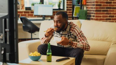 Happy guy eating noodles using chopsticks at home, opening bottle of beer and feeling relaxed at television. Smiling adult enjoying asian delivery meal for dinner, watching show on tv.